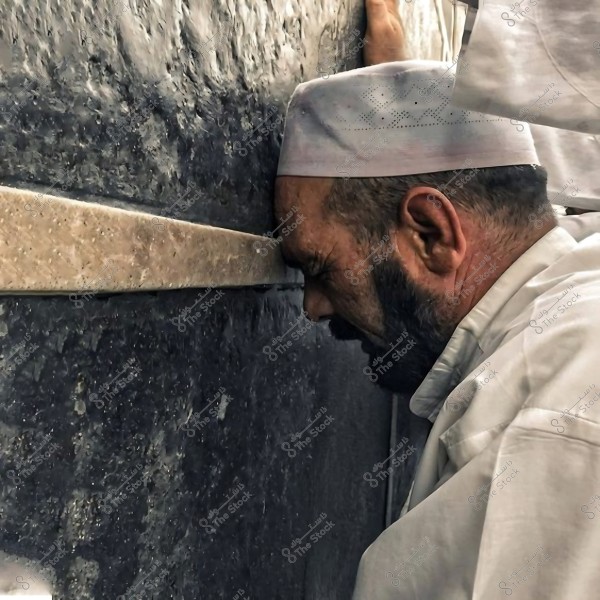 رجل يرتدي قبعة بيضاء يضع رأسه على جدار الكعبة في لحظة من الخشوع والتعبّد.