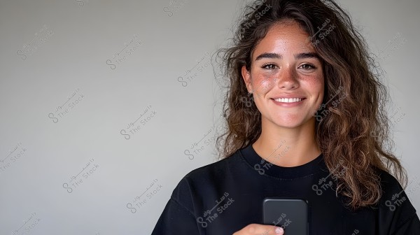 Portrait of a smiling person wearing a black shirt, holding a phone. The background is plain and monochromatic. The person has thick, curly brown hair.