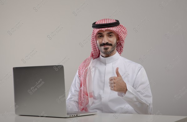 Image of a man wearing a white thobe, red checkered ghutra, and agal, sitting in front of a laptop. He appears happy and is giving a thumbs-up gesture in an indoor setting. The clothing indicates traditional Saudi attire.