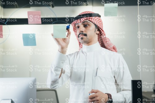 موظف سعودي يرتدي ثوب ويكتب ملاحظات العمل ويثبتها على الزجاج.