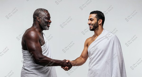 The image shows two smiling men shaking hands. Both are wearing a white ihram, a garment associated with the Hajj and Umrah pilgrimages in Islam. This attire suggests that the men might be from the Middle East or other Islamic countries. The background is plain white.