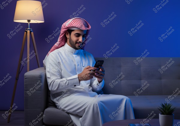 The image shows a man wearing traditional Saudi attire with a red checkered headscarf, seated on a gray couch in a room with soft blue lighting. He is focused on his mobile phone in his hands, with a small table beside him featuring a decorative plant. In the background, there\'s a lit floor lamp providing a warm ambiance to the room.