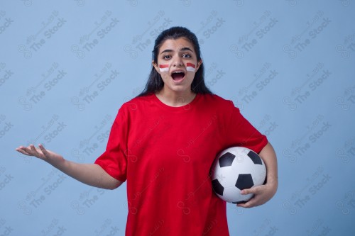 Portrait of a Saudi woman wearing a red T-shirt holding a ball, cheering the football team on a blue background and showing expressions of sadness, World Cup.