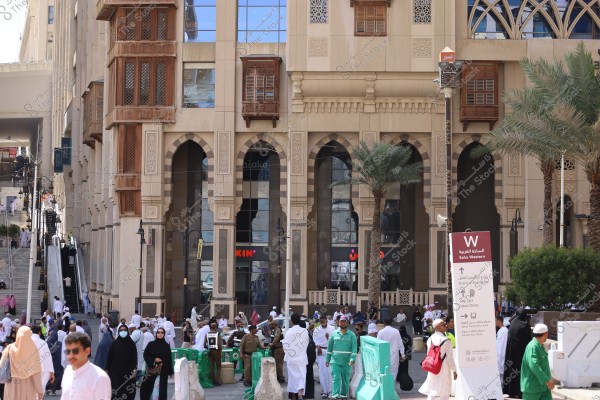 The image shows a group of people on the street near a building with Islamic architectural style featuring wooden mashrabiya windows and distinctive arches. Some individuals are wearing traditional white clothing, and there are men in green uniforms. A sign reads \"Saha Western\" with names of local streets and areas. Palm trees are also visible in the scene.