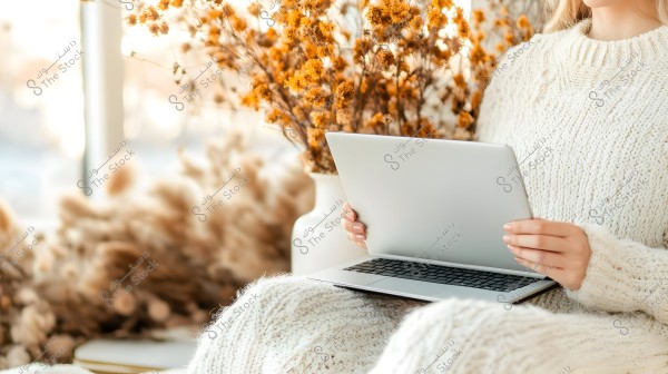 A woman sitting and wearing a cozy white knitted sweater, holding an open laptop in her hand. The background features a large vase with dried golden flowers, creating a warm and comfortable atmosphere in a room filled with natural light.