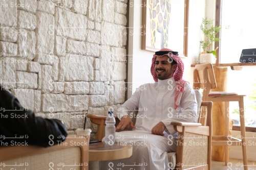 رجل سعودي يرتدي الزي السعودي التقليدي يجلس مبتسمًا ويقضي وقتًا ممتعًا في المقهى، شاب يتجاذب أطرف الحديث