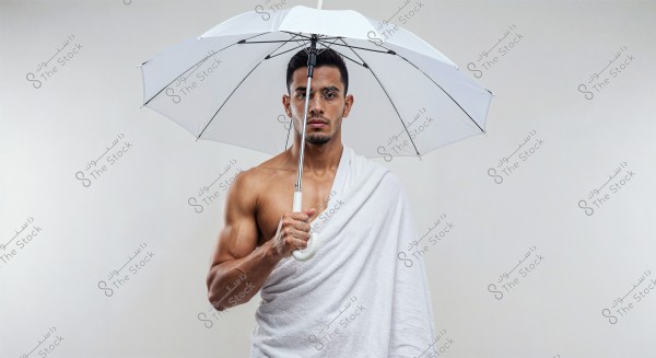 A portrait of a man standing against a white background holding a white umbrella. He is draped in a white cloth resembling an ihram. His expression is serious, and he appears to be in a traditional pose.