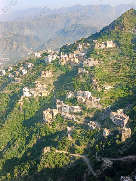 A scenic view of a mountain village in a hilly area, featuring several homes built on green terraces along the mountain slopes. A backdrop of a hazy mountain range is visible on the horizon. The buildings are closely clustered and are in neutral colors, while dense green vegetation covers the surrounding land.