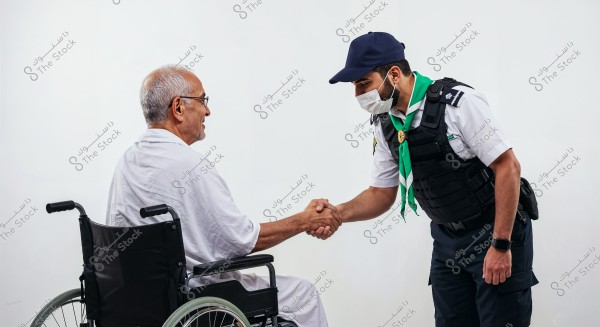 An elderly man sitting in a wheelchair wearing a white robe is shaking hands with another man dressed in a scout uniform with Saudi insignia, a blue cap, and a face mask. The standing man leans forward during the handshake.