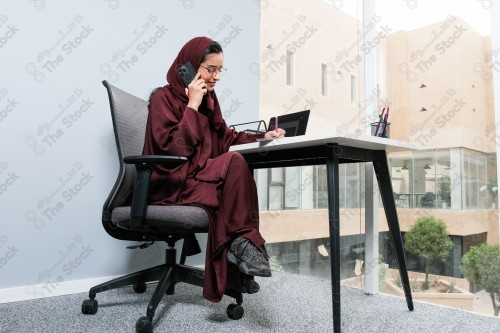 A Saudi woman wearing an abaya works in a glass-fronted office and uses a mobile device to write while taking notes during the day