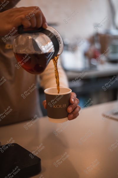 A person pouring coffee from a glass pot into a paper cup with the "KOUB" logo.