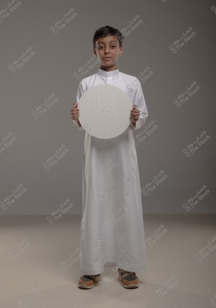 An image of a boy wearing a traditional white thobe, possibly from Saudi Arabia, standing in a dimly lit studio holding a white circular disk in his hands. The boy is also wearing brown sneakers, and the background is a neutral gray color.