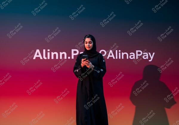 An image of a woman wearing a black abaya and a headscarf. She is standing against a gradient background transitioning from blue to pink, holding a phone in her hands. The text \"Alinn.Pay\" is displayed on the wall behind her.