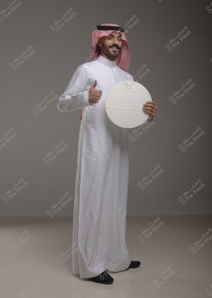 A man standing and smiling in a studio, wearing a white thobe and a red ghutra with an agal, set against a neutral gray background. He is holding a white circular board in his right hand and giving a thumbs-up with his left hand.