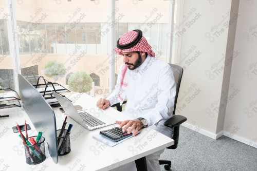 رجل سعودي يرتدي الزي السعودي التقليدي يعمل في مكتب بواجهة زجاجية ويستخدم قلم رصاص لتدوين الملاحظات