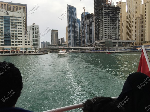 An image showcasing the skyline of Dubai with tall buildings and modern architecture. In the foreground, a boat is moving through a canal, with people seated on another partially visible boat. A red flag is visible on the boat in the lower right corner. The sky is overcast, and there is noticeable construction activity on buildings under development.