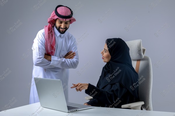 A man wearing a white thobe and red keffiyeh is standing, smiling with arms crossed. A woman in a black abaya and hijab sits on a chair, pointing at a laptop screen. The image captures a moment of discussion or collaboration in a work environment.