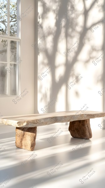 The image shows a minimalist interior design featuring a table made of heavy brown stone with an irregular surface, positioned in front of a large window allowing natural light to enter. Shadows of trees are visible on the white back wall, adding a natural and beautiful touch to the room.