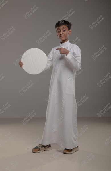 An image of a young boy wearing a traditional white thobe, standing slightly angled. In his left hand, he holds a white circle, pointing at it with his right finger. The floor is light-colored, and the background is grey.