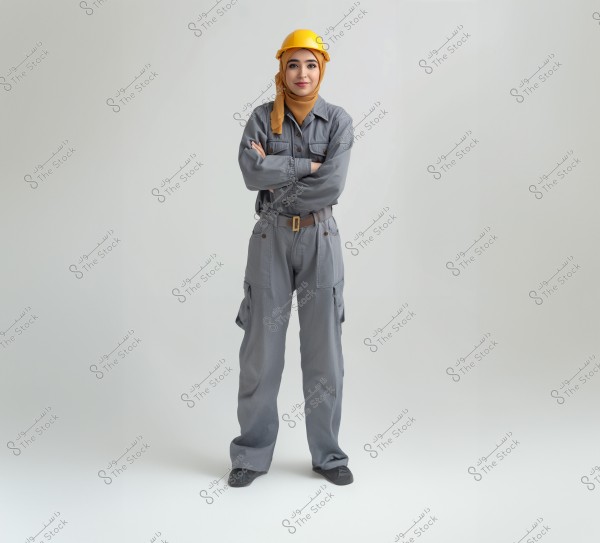 Image of a woman standing against a white background. She is wearing gray work overalls and a yellow hard hat. She appears to be of Arab or Middle Eastern descent and covers her head with a yellow hijab. Her arms are crossed on her chest, and she has a confident expression.