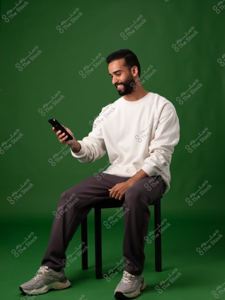 An image of a man sitting on a chair against a green background. He is wearing a white sweatshirt, gray pants, and gray sneakers. He is holding a phone and looking at it with a smile.