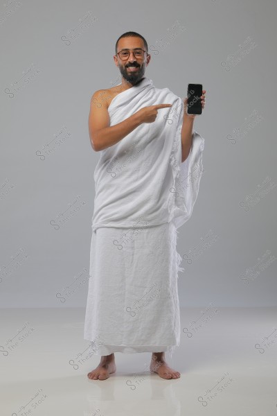 The image shows a man standing in white Ihram clothing, smiling and holding a smartphone. He is pointing to the phone with his other hand. The background is grey, with a formal and well-lit setting.