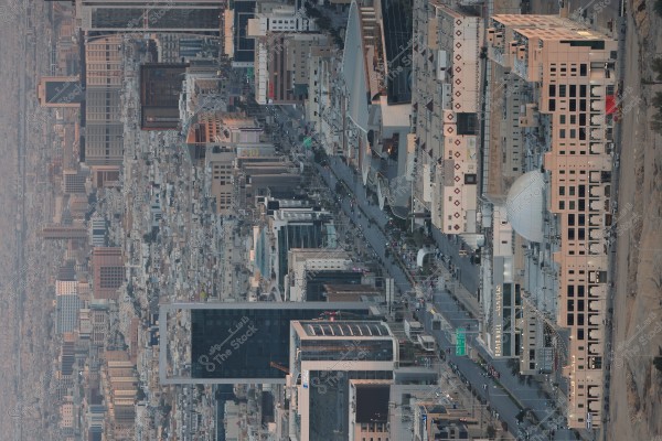 Aerial view of a modern city featuring numerous high-rise buildings and busy streets. The buildings have diverse architectural designs, including skyscrapers and glass-fronted towers. A main road bustling with vehicles and pedestrians runs through the middle. There are commercial signs on some buildings, including one with "Avenue Mall" in English and Arabic.
