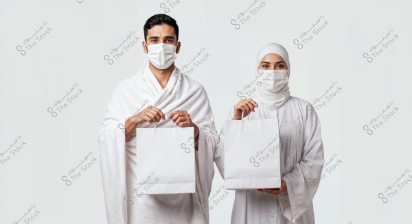 A photo of a man and a woman standing side by side, both wearing traditional white Ihram attire. The man on the left is dressed in the white izar and rida, while the woman wears a full hijab and white abaya. Both are wearing face masks and holding white paper shopping bags. The background is white.