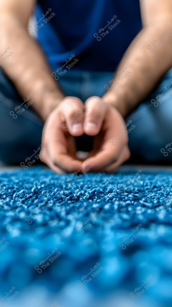 Image of a person sitting on a blue carpet, wearing a blue shirt and jeans. The focus is sharp on the blue carpet in the foreground, while the hands are clasped together in a relaxed position above the carpet.