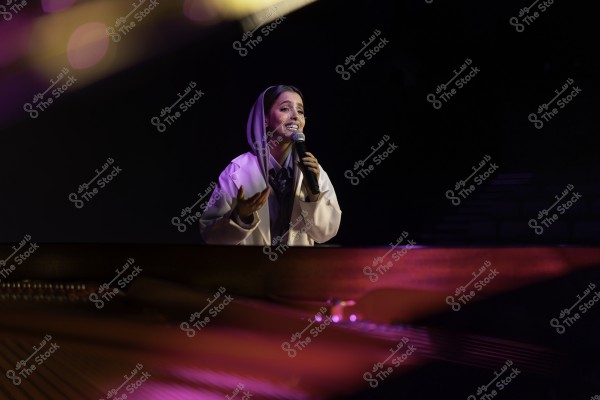 امرأة ترتدي حجابًا أبيض وتمسك ميكروفونًا وتقف على خشبة المسرح وهي تغني.