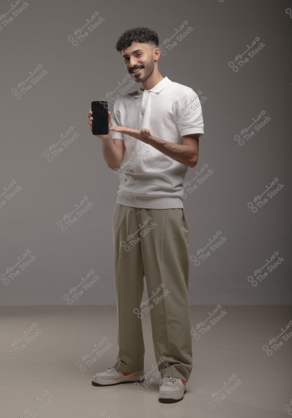 Image of a man standing in a dimly lit studio with a gray background, wearing a white polo shirt, beige pants, and white sneakers. He is holding a smartphone in one hand and gesturing towards it with the other, smiling at the camera.