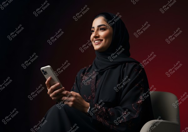 A portrait of a woman sitting on a chair holding a smartphone. She appears smiling and looking forward. She is wearing a black abaya with shiny patterns and a hijab covering her hair. The background is dark with red gradients, highlighting her facial features and attire.