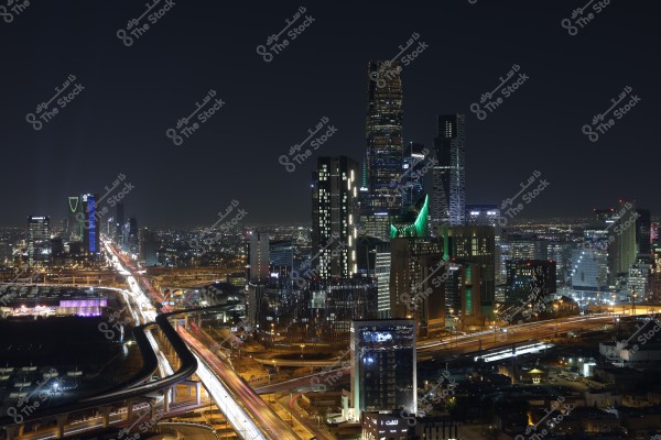 A nighttime view of Riyadh, Saudi Arabia, showcasing several skyscrapers and tall buildings. The scene is highlighted by the Kingdom Tower with its distinctive green lighting. The streets are lit by moving car lights, adding vibrancy to the urban nightscape.