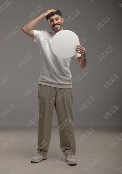 A portrait of a man standing and smiling in a gray studio, wearing a white shirt, beige pants, and sneakers. He holds a white circle in his right hand while placing his left hand on his head.