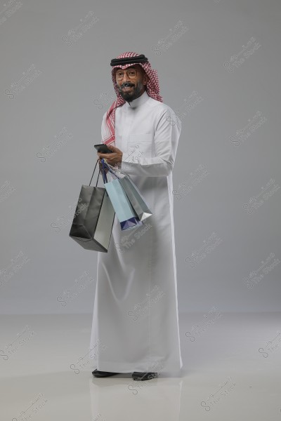 A man wearing traditional Saudi attire with a red headscarf and glasses, holding several shopping bags of different colors and a mobile phone. He looks happy and stands in a neutral studio setting with a light gray background.