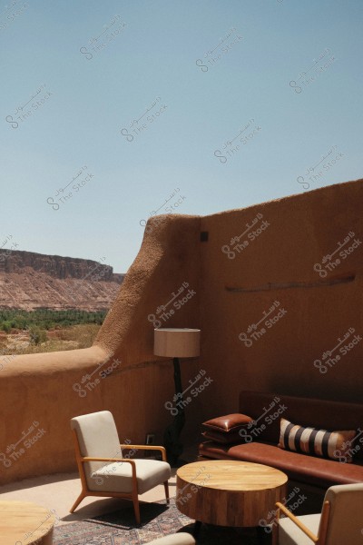 The image shows an outdoor seating area featuring a brown sofa, two light-colored armchairs, and a round wooden coffee table. There is also a floor lamp. The walls and floor are clay-colored, with a backdrop of rocky mountains and green vegetation under a clear blue sky.