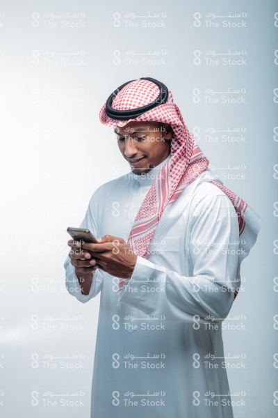 شاب سعودي يرتدي ثوب أبيض على خلفية بيضاء ، يمسك الجوال ، يستخدم أحد تطبيقات الآيفون ، يتصفح الإنترنت ، يستخدم وسائل التواصل الاجتماعي