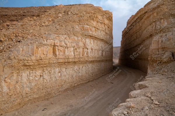 The image shows a narrow canyon in a desert area between high rock walls. The rocks have distinct layers in various shades of brown and beige. The sky is blue with some white clouds visible in the background. The ground is covered with dust and sand.