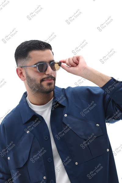 Image of a man wearing round sunglasses adjusted with one hand, with neatly styled black hair. He is dressed in a dark blue shirt over a white T-shirt, set against a bright white background. His expression is relaxed and friendly.
