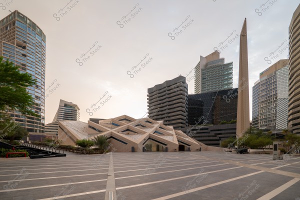 A spacious plaza in front of a collection of modern buildings and skyscrapers with various architectural designs. A uniquely designed geometric building stands in the center with a tall obelisk beside it. Trees line the sides, and the sky is visible in the background with soft natural lighting.