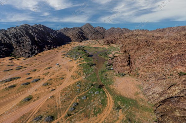 A beautiful natural landscape showing a valley between a range of rocky mountains. The ground is covered with sand and rocky terrain with patches of green vegetation. There are several dirt tracks winding through the scene. The sky is blue with scattered white clouds.