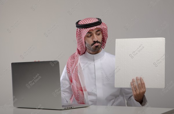 A portrait of a man sitting in front of a laptop wearing traditional Saudi attire, a white thobe, red and white checkered ghutra, and agal. He is holding a blank white board in his right hand and looking at it.