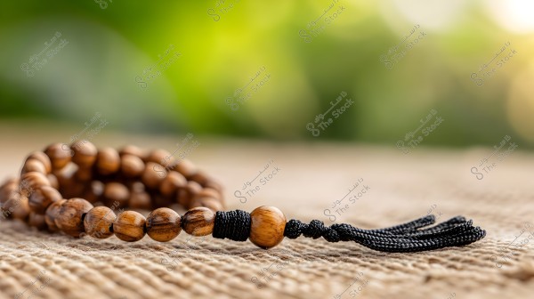 A close-up image of a prayer bead string made from polished wooden beads, featuring a sturdy black thread ending in a knot and tassels. The prayer beads are placed on a burlap surface, with a blurred green background adding a sense of calmness and focus to the beads.