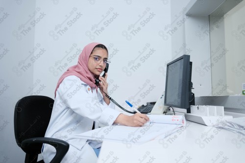 Saudi doctor wearing a medical uniform, sitting in her office and taking notes, the health field, medical consultations and medical health services.