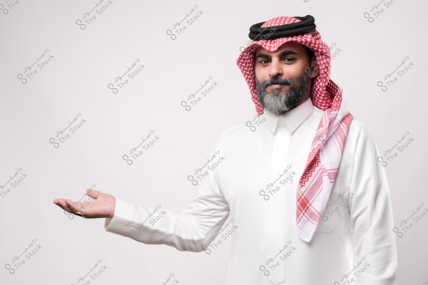 A man wearing traditional Gulf attire, including a white thobe, red checkered headscarf, and agal, gesturing welcomingly with his hand.