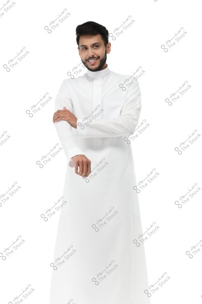 A portrait of a smiling man wearing a traditional white thobe, standing facing the camera against a white background. The man is adjusting the sleeve of the thobe with his left hand. The attire suggests the man might be from the Gulf region.