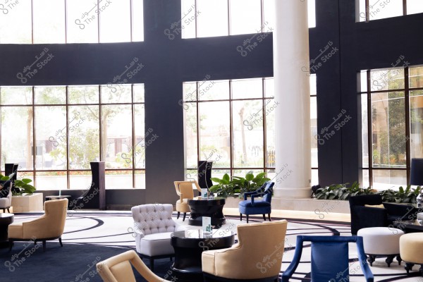 The image depicts a large indoor seating area with modern and elegant decor. The space features large glass windows allowing plenty of natural light and views of trees and plants outside. Inside, there are chairs in various colors like beige and blue, and small tables in between. Green plants adorn the area, adding a touch of nature to the interior design.