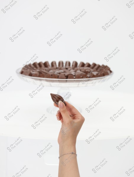 A hand holding an oval-shaped chocolate piece, with a glass fruit tray filled with similar chocolate pieces arranged in a circular pattern in the background. The background is white and clean, giving the image a sense of purity and elegance.
