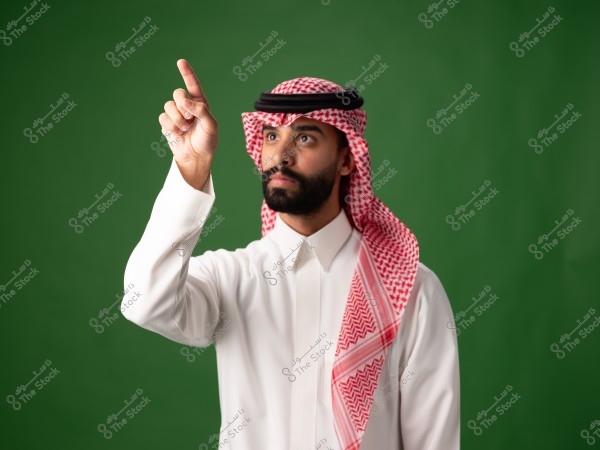 Portrait of a man wearing traditional Saudi attire, consisting of a white thobe, red checkered headscarf (ghutra), and black headband (agal), standing against a green background. The man is pointing upwards with his index finger.