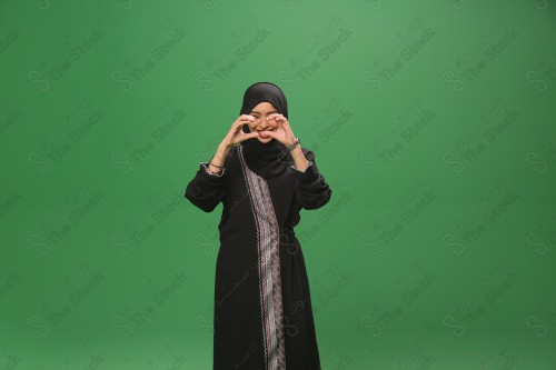 Saudi woman on green background wearing abaya and hijab, making gestures with her hands, making a heart with her fingers while smiling, Eid celebrations